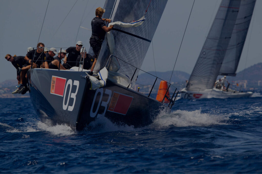 This image shows a competitive sailing scene with a team of crew members actively engaged in maneuvering a sailboat. The boat, bearing the number 03, is heeling over in the water indicating speed and the force of the wind on its sails. The crew members are positioned strategically to balance the boat, with some sitting on the edge and one standing, likely adjusting the sail. The background is a deep blue sea with waves, and there's another sailboat with a number 05 in the distance, suggesting a race or regatta. The focus and coordination of the crew highlight the intensity and teamwork required in the sport. The technology and materials used for the boat's hull and sails might be of particular interest to a composite material manufacturer, as they demonstrate the application of high-performance materials in marine environments.